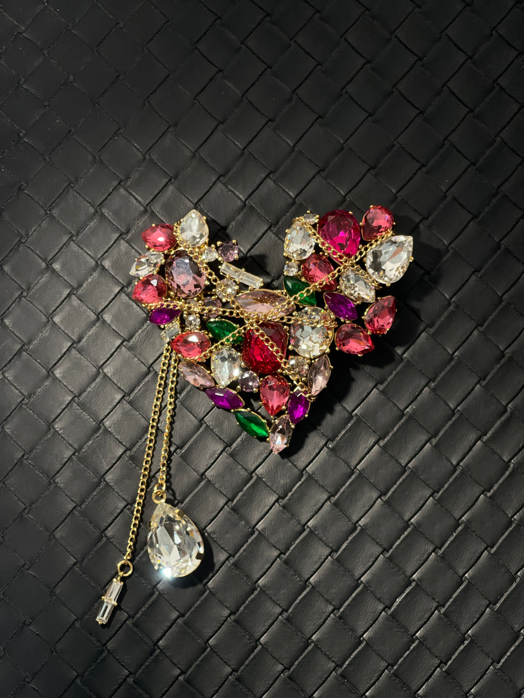 Heart-shaped gold-tone brooch with multicoloured faceted crystal stones and decorative chain tassels on black woven leather background.