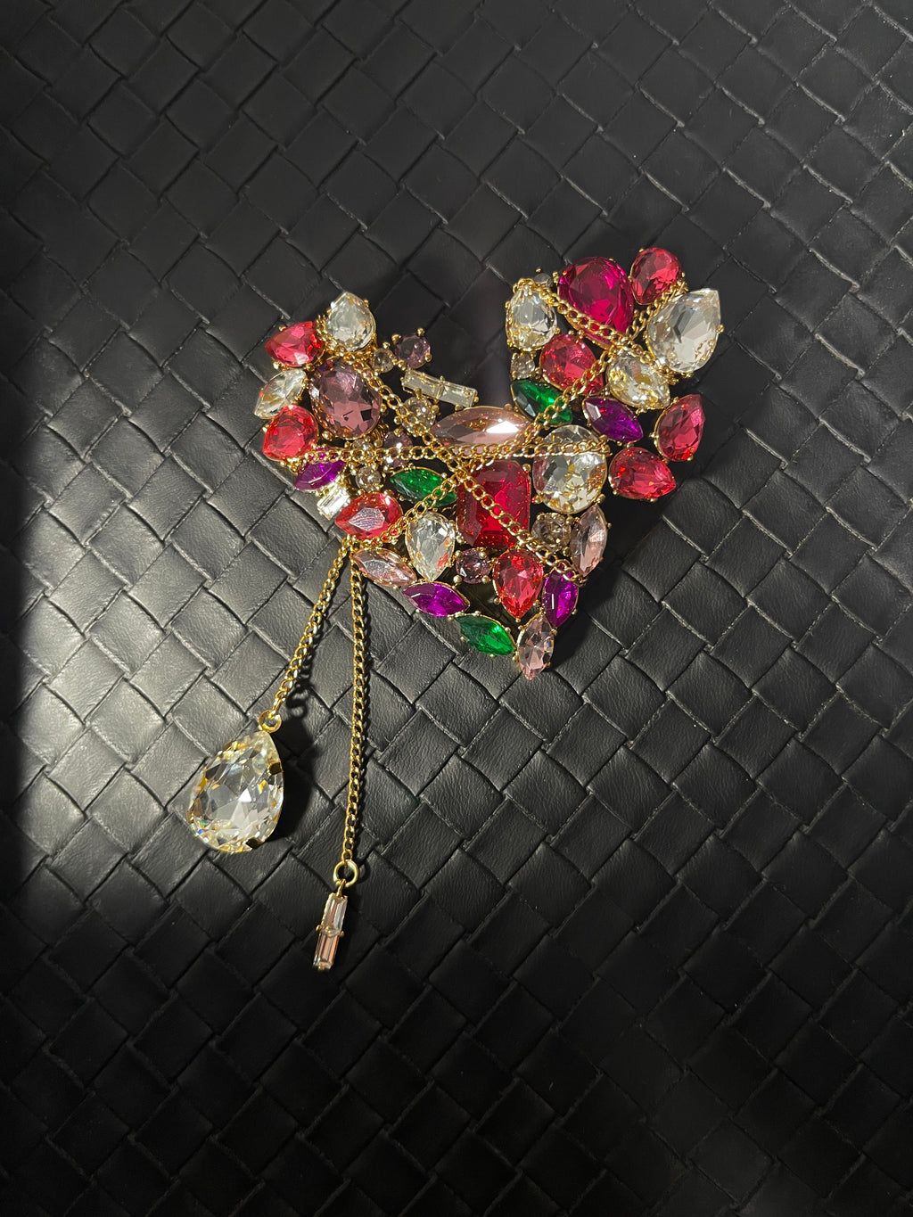 Gold-tone heart brooch with colourful faceted stones, decorative chains and crystal drop tassels on textured black leather surface.